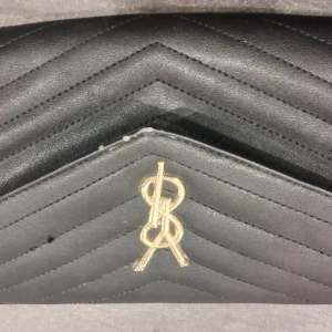 Svart YSL kuvertväska med kedja - Snygg svart kuvertväska från YSL i skinn med quiltad design och guldiga detaljer. Väskan har en elegant kedjerem och klassisk YSL-logga framtill. Magnetlås och innerfack med dragkedja gör den både praktisk och stilren.