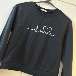 Svart sweatshirt med hjärtpuls-tryck - Svart sweatshirt med rund hals och lång ärm. Framtill finns ett vitt tryck med pulslinje och hjärta. Tröjan har ribbade muddar vid ärmslut och nederkant. Perfekt för en chill och stilren look.