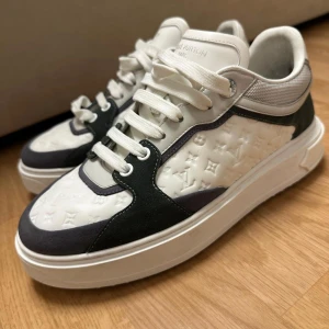 Louis Vuitton vita och svarta sneakers - Snygga Louis Vuitton sneakers i vitt och svart med präglat LV-mönster på sidorna. Skorna har tjock vit sula, snörning och detaljer i mocka och skinn. Silverdetaljer på hälen och meshpartier på sidan ger en lyxig och modern vibe.