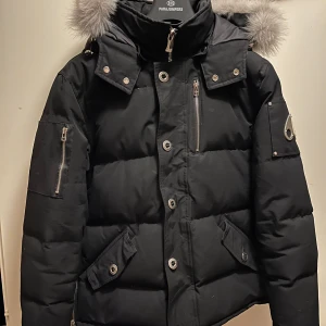 Svart dunjacka Moose Knuckles L - Säljer en svart dunjacka från Moose Knuckles i storlek L. Jackan har pufferdesign, silverfärgade detaljer, dragkedjor och knappar, samt en stor huva med fluffig päls. Klassisk logga på ärmen och flera praktiska fickor. Perfekt för kalla dagar. (Edit) GAMLA MODELLEN MED ÄKTA PÄLS, de nya modellerna görs inte längre med äkta päls därav är den här lite dyrare.