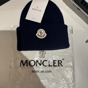 Mörkblå ribbstickad mössa Moncler - Snygg mörkblå ribbstickad mössa från Moncler med klassisk uppvikt kant och broderad logotyp framtill. Perfekt accessoar för kyliga dagar, tillverkad i mjukt och värmande material. En stilren och tidlös mössa som lyfter vilken outfit som helst.