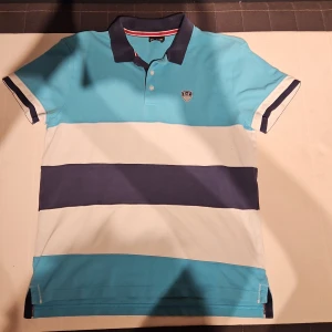 Randig EA7 Emporio Armani pikétröja - Snygg pikétröja från EA7 Emporio Armani med breda ränder i turkos, vitt och marinblått. Klassisk krage, knappar framtill och logga på bröstet. Perfekt för dig som gillar stilren och sportig look. Man kan se lite slitage på logga som är på bröstet.