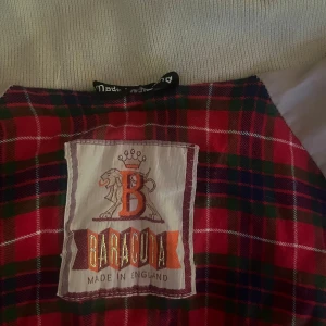 Beige Baracuta bomberjacka med rutig insida - Säljer en klassisk beige bomberjacka från Baracuta, tillverkad i England. Jackan har en snygg röd-rutig insida, ribbade muddar och krage med knapp. Två fickor framtill med knappstängning och dragkedja. Perfekt för dig som gillar tidlös stil med brittisk touch.