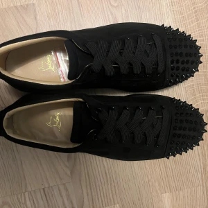 Christian Louboutin svarta sneakers med nitar - Svarta sneakers från Christian Louboutin i mocka med coola svarta nitar på tån. Skorna har rund tå, platt sula och klassisk snörning. Insidan är beige och loggan syns på innersulan. Perfekta för dig som vill sticka ut med en edgy vibe.
