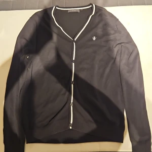 Svart kofta från Riley med vita detaljer - Svart kofta från Riley i storlek M med vita ränder längs knäppningen och halsringningen. Koftan har knappar framtill, lång ärm och en broderad logga på bröstet. Perfekt för lager-på-lager och stilrena outfits. Perfekt om man vill vara snyggt klädd på vintern.