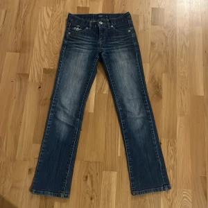 Blå 2000s jeans - Snygga blå jeans från Comp. by KappAhl i storlek 146. Modellen har bootcut-ben, klassisk femficksdesign och coola slitningar framtill. Jeansen har broderad detalj på fickan och knappar med logga. Perfekta för dig som gillar en cool stil! Anndvända få gånger