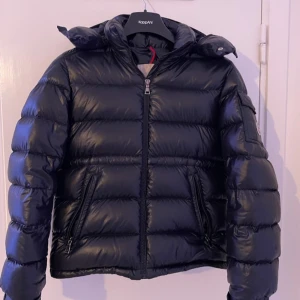 Moncler maya  - Säljer en svart pufferjacka från Moncler med klassisk logga på ärmen. Jackan har hög krage, huva och dragkedja framtill samt två fickor med dragkedja. Fyllig och glansig yta som ger en riktigt snygg look. Perfekt för kalla dagar.