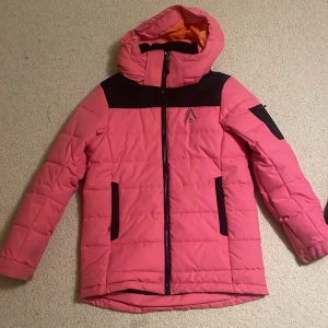 Rosa pufferjacka med huva - Snygg rosa pufferjacka från Everest med svarta detaljer och huva. Jackan har dragkedja framtill, två svarta fickor med dragkedja och en svart detalj på ena ärmen. Perfekt för kalla dagar och har en sportig vibe. Storlek 150, men känns som en typ s 