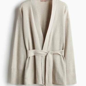 Beige kofta H&M - Snygg beige kofta med omlott-design och knytbälte i midjan. Koftan har långa ärmar med breda ribbade muddar och en mjuk, avslappnad passform. Perfekt för att slänga på sig när det är lite kyligt ute.