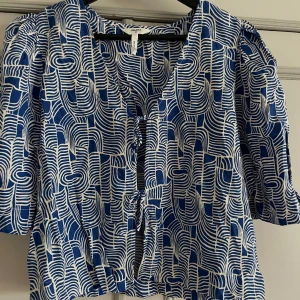 Blåvit blus med knyt från Object - Säljer en blåvit blus från Object i storlek 38. Blusen har ett grafiskt mönster, korta puffärmar och snygga knytningar framtill. V-ringad och luftig modell som ger en cool och kreativ vibe. Perfekt för dig som gillar unika detaljer och mönster.
