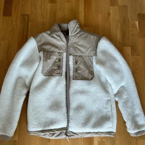Beige teddyjacka från H&M - Säljer en beige och vit teddyjacka från H&M med två stora bröstfickor och dragkedja framtill. Jackan har kontrasterande partier i beige nylon och mjuk teddyfleece ny pris 700