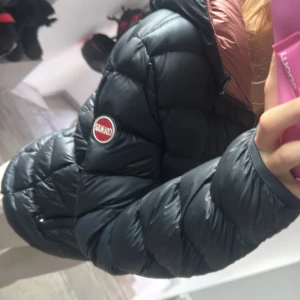 Blå pufferjacka från Colmar - Säljer en blå pufferjacka från Colmar med rosa insida på huvan. Jackan har dragkedja framtill, två fickor och ett broderat Colmar-märke på ärmen. Den är quiltad och har en klassisk, sportig look som är perfekt för kyliga dagar. Den har några små hål men inget som märks