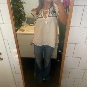 Vit blus med broderi från H&M - Vit blus från H&M med svarta broderade blommor och dekorativa band med tofsar vid halsen. Blusen har korta volangärmar och är tillverkad i bomull. Perfekt för en bohemisk look med snygga detaljer och luftig passform. Den har ett litet hål på ryggen som knappt syns men annars jätte söt🩵🩷💖✨🫰🏻