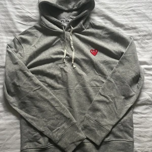 Grå hoodie Comme des Garçons Play i storlek S - Grå hoodie från Comme des Garçons i storlek S men passar även M. Tröjan är helt oanvänd tagsen borta dock. Skrov vid fler funderingar 