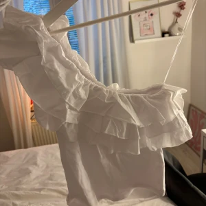 Vit oneshoulder topp med volanger - Säljer en vit oneshoulder topp med flera lager volanger längs axeln och halsringningen. Toppen har en asymmetrisk design med ett smalt axelband på ena sidan och är tillverkad i ett lätt bomullstyg från Zara. Använt 1 gång, så i princip som ny!