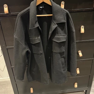 Svart overshirt från Gina Tricot - Svart overshirt från Gina Tricot i storlek M. Jackan har två stora bröstfickor med lock, klassisk krage och knäppning framtill. Materialet är mjukt och känns som ull, perfekt för lager-på-lager. Stilren och enkel design som funkar till många outfits.
