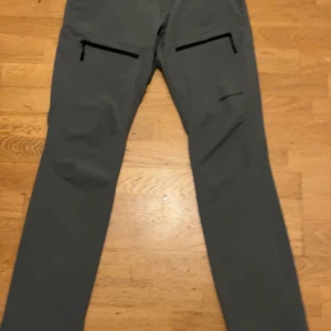 Grå täckbyxor från Trekmates, S - Snygga grå täckbyxor från Trekmates i storlek S. De har raka ben, två framfickor med dragkedja och knappstängning i midjan. Perfekta för utomhusaktiviteter och gjorda i slitstarkt syntetmaterial som står emot väder och vind.