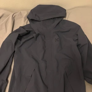 Mörkblå vindjacka från Haglöfs L - Snygg mörkblå vindjacka från Haglöfs med huva och minimalistisk design. Jackan har diskret Haglöfs-logga på bröstet och meshfoder på insidan för extra komfort. Perfekt för blåsiga dagar och outdoor-aktiviteter.