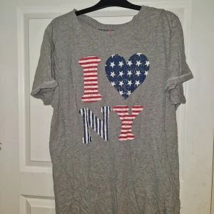 Grå I Love NY t-shirt - Säljer en grå t-shirt med trycket 'I ♥ NY' i amerikanska flaggans färger och mönster. T-shirten har korta ärmar och är tillverkad i mjuk bomull. Perfekt för dig som gillar streetstyle och vill visa din kärlek till New York.