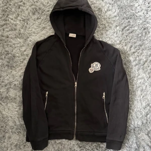 Svart Moncler hoodie med dragkedja XXL - Svart hoodie från Moncler i storlek XXL med huva och hel dragkedja framtill. Tröjan har två fickor med dragkedja och ett snyggt Moncler-märke på bröstet. Tillverkad i mjuk bomull, perfekt för chill dagar. Långärmad och stilren design.