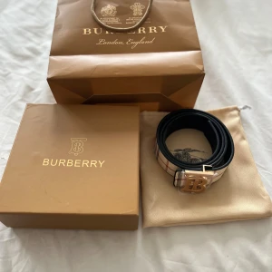 Burberry bälte - Säljer ett snyggt Burberry bälte med klassiskt rutigt mönster i beige, svart och rött. Bältet har ett stort guldigt 'B'-spänne och är tillverkat i skinn med textil på framsidan. Perfekt accessoar för dig som vill sticka ut med lyxig stil.