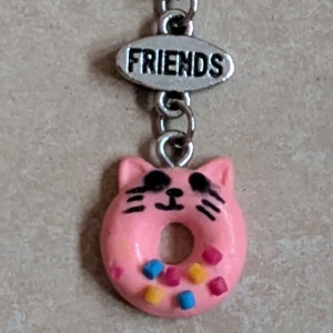 Claire's halsband med katt-donut - Supergulligt halsband från Claire's med en silverfärgad kedja och ett hänge som föreställer en rosa donut i form av en katt, dekorerad med färgglada strössel. Ovanför hänget sitter en liten bricka med texten 'FRIENDS'. Perfekt accessoar för dig som älskar söta detaljer.