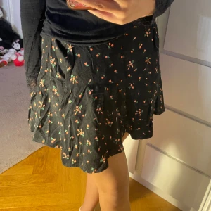 Blommig omlott kjol - Supersöt svart kjol med små blommor från Urban Outfitters 🌷 Lätt och flowy tyg – perfekt till både vardag och sommaroutfits. Storlek: M men passar även S Fint skick  🖤 Passar perfekt med en enkel topp eller stickad tröja!