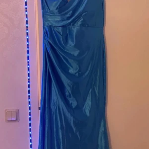 Blå långklänning med oneshoulder - Säljer en snygg blå långklänning med glansigt tyg och oneshoulder-design. Klänningen har draperade detaljer framtill och en axelbandslös sida som ger en cool och modern look. Perfekt för dig som vill sticka ut med en unik stil.