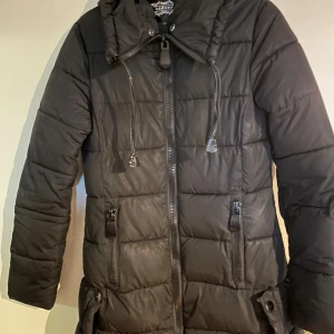 Svart lång pufferjacka från Garoff - Svart lång pufferjacka från Garoff med huva, dragkedja framtill och två stora fickor med dragkedja. Jackan har quiltad design och justerbar dragsko vid halsen. Perfekt för kalla dagar när du vill hålla dig varm och snygg.