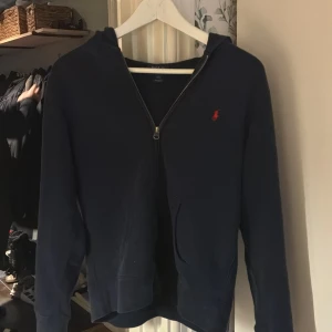 Marinblå hoodie från Polo Ralph Lauren - Snygg marinblå hoodie från Polo Ralph Lauren med röd broderad logga på bröstet. Tröjan har dragkedja framtill, huva och två fickor. Perfekt för chill dagar och har en klassisk look som funkar året runt.