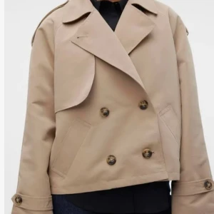 Beige kort trenchcoat från Vero Moda - Kort trenchcoat i beige från Vero Moda med dubbelknäppning, bred krage och snygga knappar på ärmslut och axlar. 