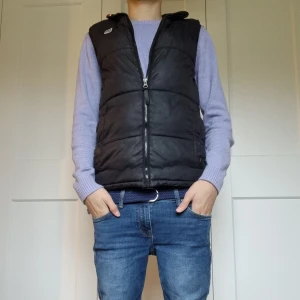 Svart dunväst från Lee med dragkedja - Svart dunväst från Lee med quiltad design och hel dragkedja framtill. Västen har en stilren look med diskret logga på bröstet och två sidofickor. Perfekt att slänga på över en hoodie eller tröja för extra värme. Passformen är normal och materialet känns mjukt och lätt. Modellen på bilden är 171 cm lång men skulle säga att den passar från 160 till 170