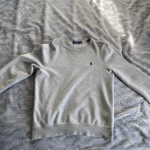 Grå sweatshirt från Polo Ralph Lauren - Klassisk grå sweatshirt från Polo Ralph Lauren med rund halsringning och broderad logga på bröstet. Tröjan har ribbade muddar vid ärmslut och nederkant, samt en snygg detaljsöm vid halsen. Perfekt för en chill och stilren look. Skick 9/10 och qr koden är sliten men fungerade förut!