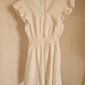 Ljus kortklänning med volangärmar - Supersöt ljus kortklänning med v-ringning, volangärmar och smock i midjan. Klänningen har diskret struktur i tyget och spetsdetaljer längs ringningen. Perfekt för sommarens alla tillfällen!