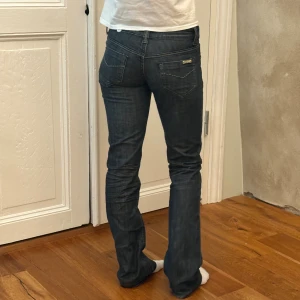 Mörkgrå bootcut jeans - Säljer ett par mörkblå/grå jeans  med klassisk femficksdesign och diskreta kontrastsömmar. Modellen har låg midja och bootcut-ben, vilket ger en snygg siluett. Jeansen är tillverkade i stretchig denim för extra komfort och har en metallknapp framtill. Midjemått mått 36
