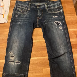 Replay Anbass jeans med slitningar - Snygga mörkblå Replay Anbass jeans med coola slitningar och distressed detaljer på både fram- och baksida. Klassisk femficksmodell med kontrastsömmar och broderade bakfickor. Perfekt passform och grym streetkänsla. 