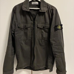 Grå/grön overshirt från Stone Island - Säljer en svart overshirt från Stone Island med klassisk patch på ärmen. Jackan har två bröstfickor med knappar, dragkedja framtill och en rak passform. Tillverkad i bomull, perfekt för lager-på-lager och streetwear-stil.