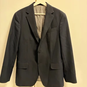 Mörkblå kavaj från Oscar Jacobson - Klassisk mörkblå kavaj från Oscar Jacobson med två knappar framtill och fickor med lock. Fodret har ett stilrent rutigt mönster i svart och vitt. Perfekt för en dressad look och snygg passform.