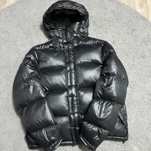 Svart pufferjacka Emporio Armani - Snygg svart pufferjacka från Emporio Armani med glansig finish och huva. Jackan har dragkedja, snörning vid huvan och diskret logga på bröstet. Insidan är olivgrön och jackan är tillverkad i lätt polyester. Perfekt för kalla dagar när du vill hålla stilen.