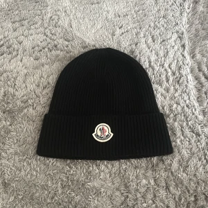 Svart Moncler mössa - Svart ribbstickad mössa från Moncler med uppvikt kant och klassisk Moncler-logga framtill. Perfekt för att hålla värmen och ge en clean look. Materialet är mjukt och stretchigt, vilket gör den bekväm att bära hela dagen.