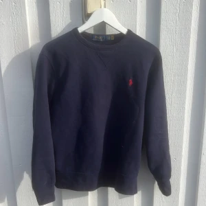 Mörkblå sweatshirt från Polo Ralph Lauren - Klassisk mörkblå sweatshirt från Polo Ralph Lauren med röd broderad logga på bröstet. Tröjan har rund hals, långa ärmar och ribbade muddar vid ärmslut och nederkant. Perfekt för en chill och stilren look.