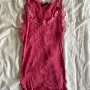 Rosa ribbad linnetopp från Rosemunde - Supersöt rosa linnetopp från Rosemunde med ribbad struktur och spetsdetaljer runt halsringningen och axlarna. Modellen är ärmlös och har en feminin v-ringad form. Perfekt att styla med jeans eller kjol för en chill look.