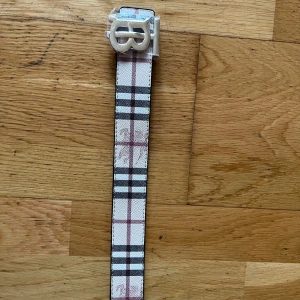 Burberry rutigt bälte med silverdetalj - Säljer ett snyggt Burberry bälte med klassiskt rutigt mönster i beige, svart, vit och röd. Bältet har en stilren silverfärgad spänne med Burberrys logga. Materialet är skinn och bältet kommer med originalpåse och Burberry shoppingbag. Man får med en grej som man kan göra hål med i bältet. Bältet är dubbel sidig.