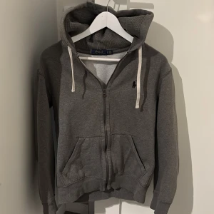 Grå hoodie från Polo Ralph Lauren - Snygg grå hoodie från Polo Ralph Lauren med dragkedja, huva och vita snören. Klassisk logga på bröstet och två fickor framtill. Perfekt för chill dagar och enkel att matcha med jeans eller joggers.