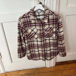 Rutig overshirt från Lindex Kids - Snygg rutig overshirt från Lindex Kids i beige, rosa och svart. Skjortan har långa ärmar, klassisk krage, två bröstfickor med knappar och ett mjukt material som känns skönt mot huden. Perfekt för lager-på-lager och chill stil.