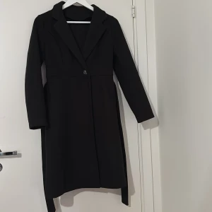 Svart kappa med bälte och knappar - Stilren svart kappa med klassisk krage, knäppning framtill och tillhörande bälte i midjan. Långärmad modell med rak passform och snyggt fall. Perfekt för dig som gillar en clean och tidlös look.