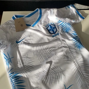Vit Brasilien fotbollströja Nike - Snygg vit Brasilien fotbollströja från Nike med blå detaljer och palmbladsmönster. Tröjan har korta ärmar och rund halsringning med blå kant. Perfekt för dig som vill sticka ut på planen eller heja på landslaget.