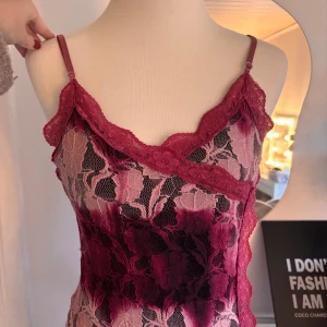 Rosa spetstopp vintage med blommönster - Supersöt kort klänning i rosa och vinröda toner med blommig spets och tunna axelband. Klänningen har en figurnära passform och v-ringad hals med dekorativ spetskant. 