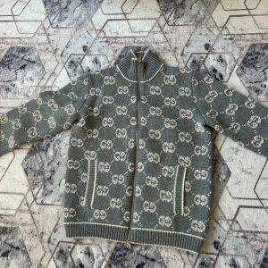 Grå kofta från Gucci i ull - Exklusiv grå kofta från Gucci i 100% ull med hel dragkedja och ribbade muddar. Tröjan har ett snyggt mönster med Guccis klassiska GG-logga i vitt över hela plagget och två fickor framtill. Perfekt för dig som vill sticka ut med lyxig streetstyle.
