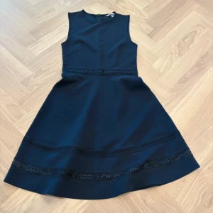 Marinblå ärmlös klänning från H&M - Snygg marinblå klänning från H&M med ärmlös design och rund hals. Kjolen är utsvängd och har dekorativa spetsdetaljer med små cirklar vid midjan och nedtill. Klänningen har en diskret dragkedja i ryggen och är tillverkad i ett strukturerat polyestertyg.